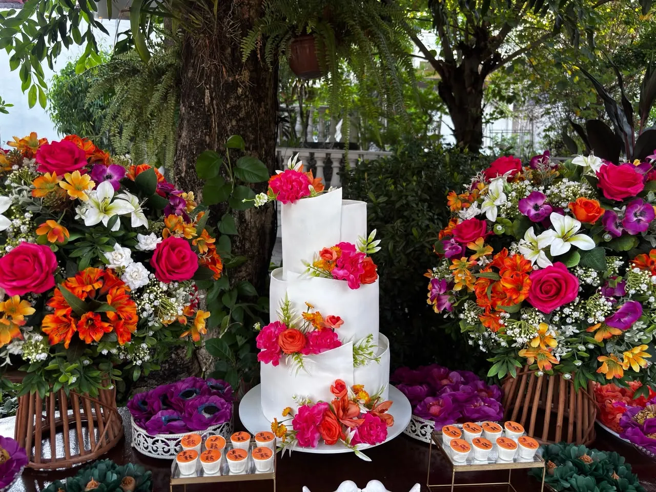 Bolo branco decorado com flores entre grandes arranjos coloridos