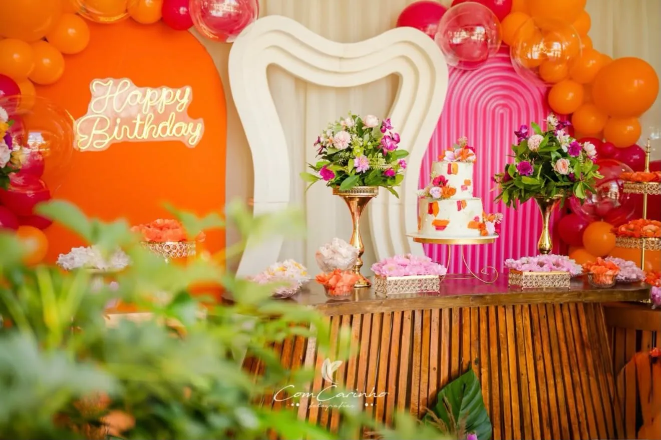 Mesa de aniversário com bolo, flores e balões laranja e rosa