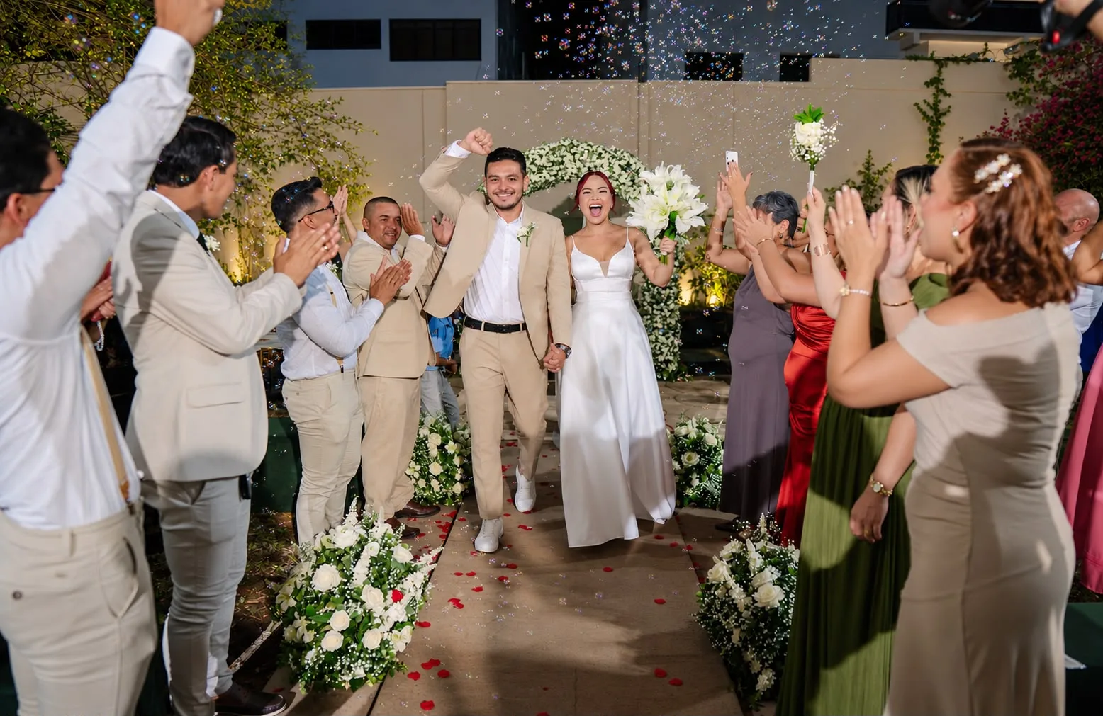 Casal entrando na celebração entre pétalas, confetes e convidados aplaudindo