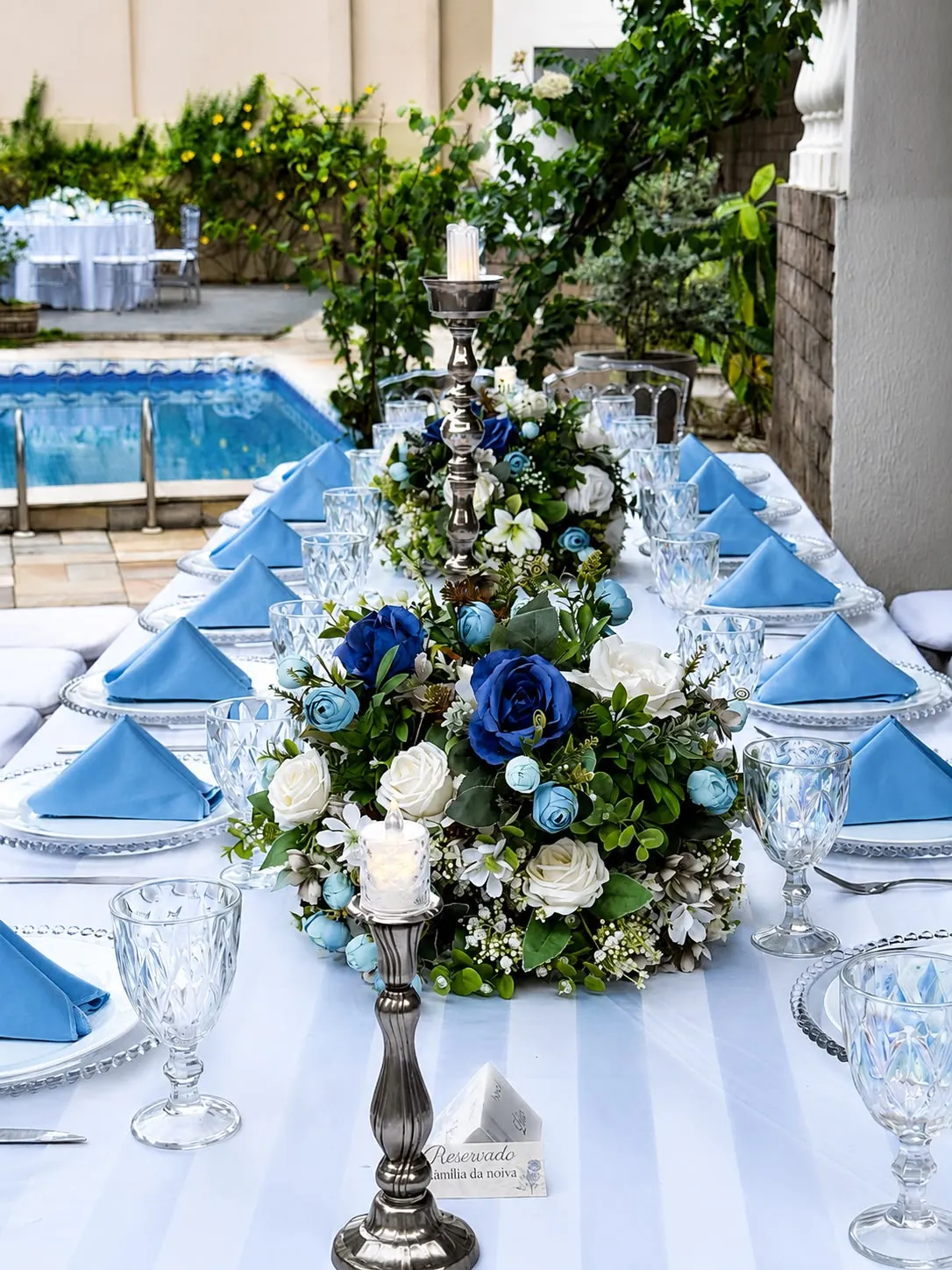 Mesa posta com guardanapos azuis, taças de cristal e arranjos de rosas ao lado da piscina