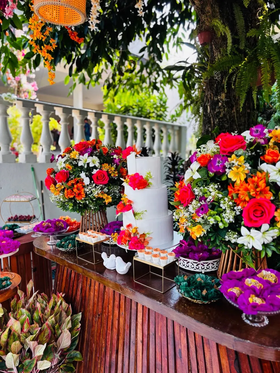 Mesa de bolo branco com arranjos de flores coloridas sob as árvores do jardim