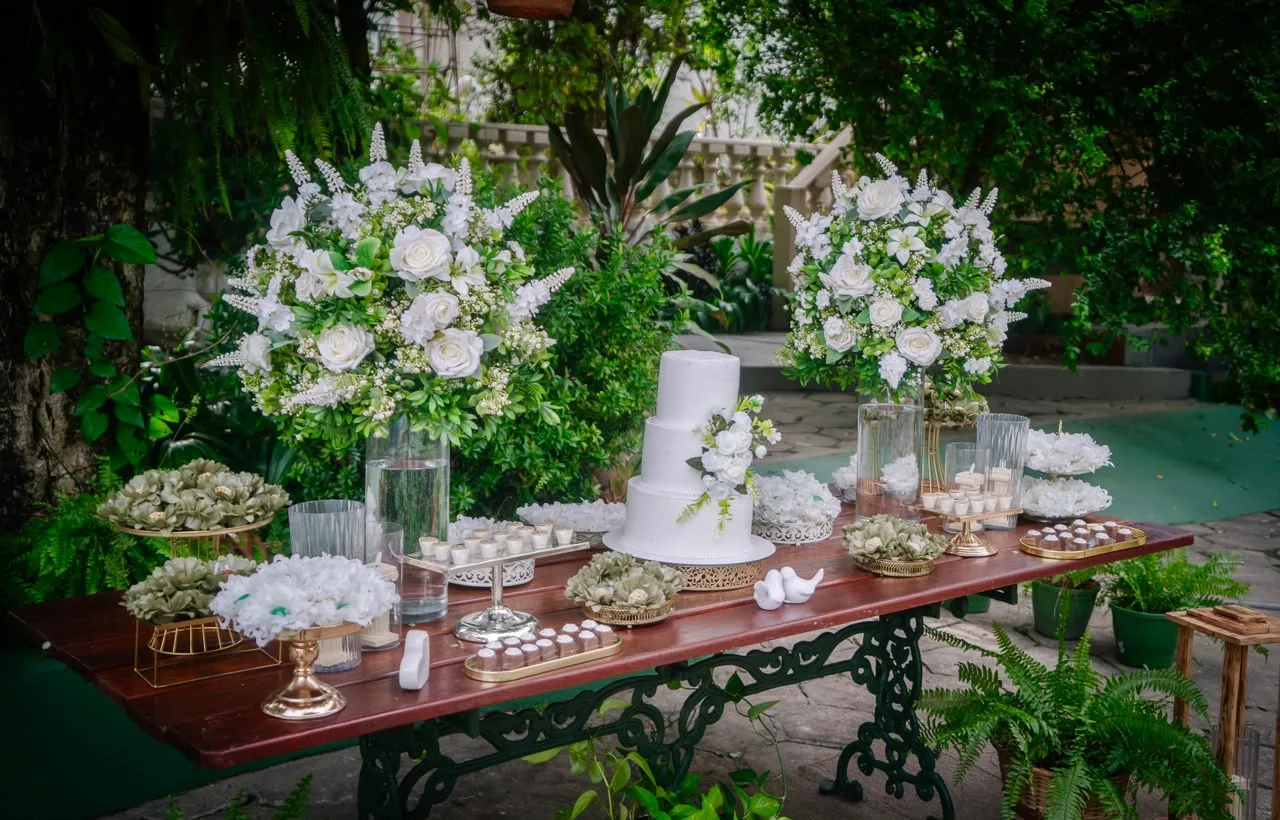 Mesa de doces com bolo branco e arranjos de flores brancas no jardim